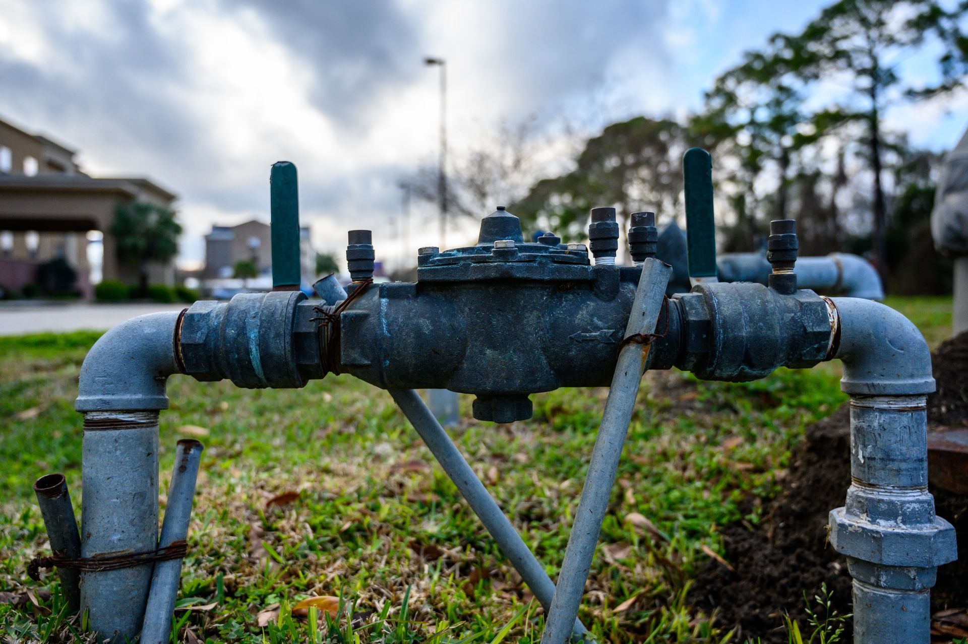 Weathered metal pipeline valve in a grassy yard, with houses and cloudy sky in the background.