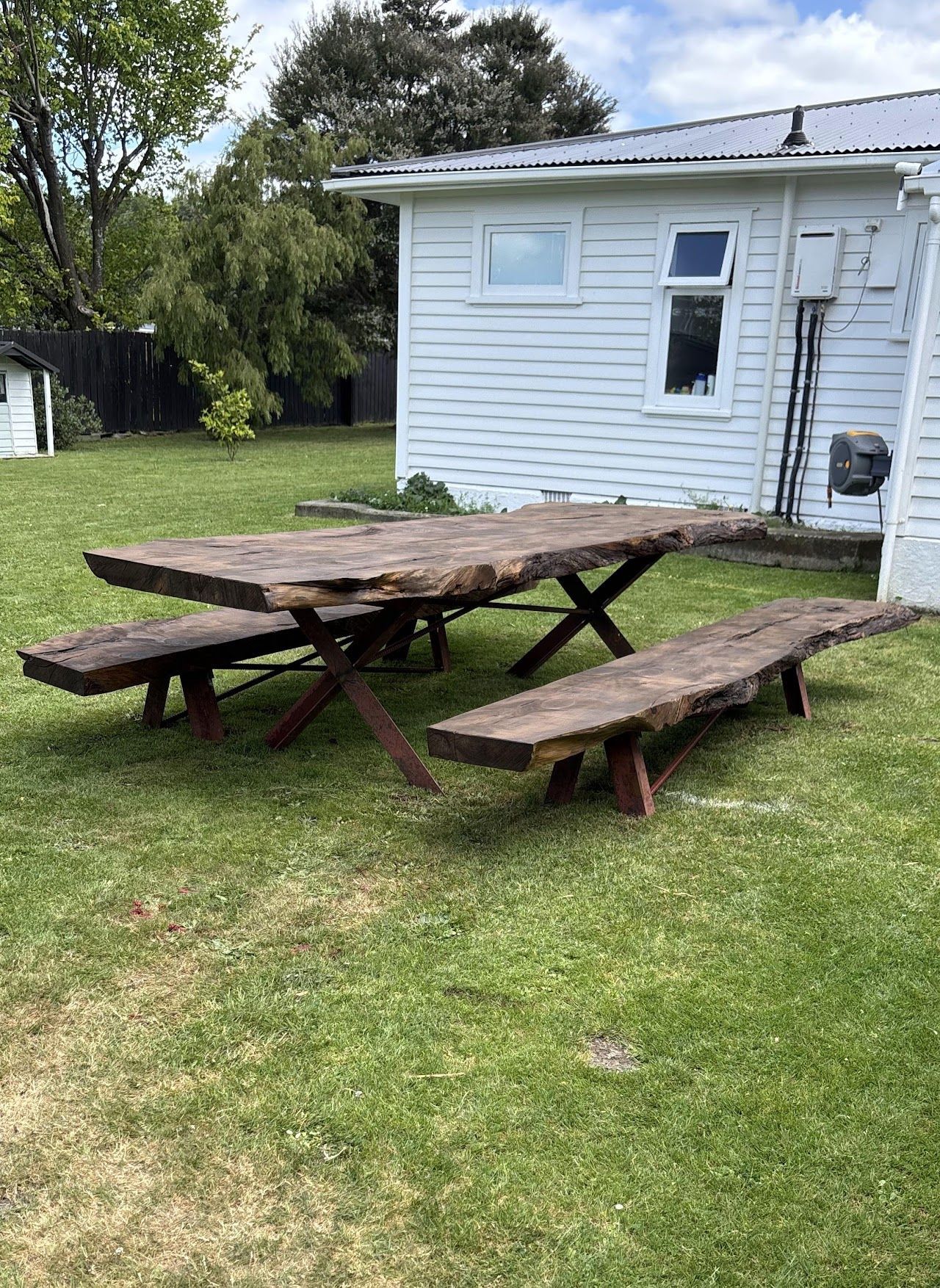 A wooden picnic table and benches are sitting in the grass in front of a white house.
