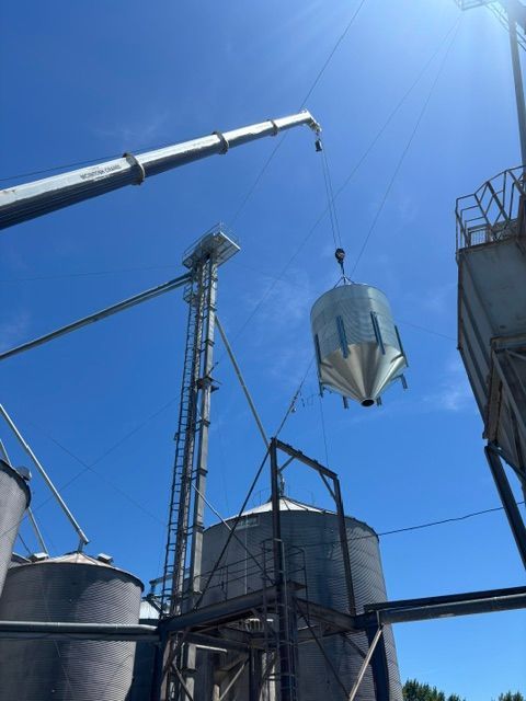 Crane lifting a metal grain hopper; other silos and a ladder visible against a blue sky.