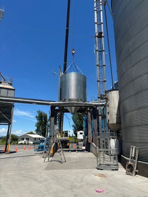 Crane lifting a large, cylindrical metal component into a grain silo structure on a sunny day.
