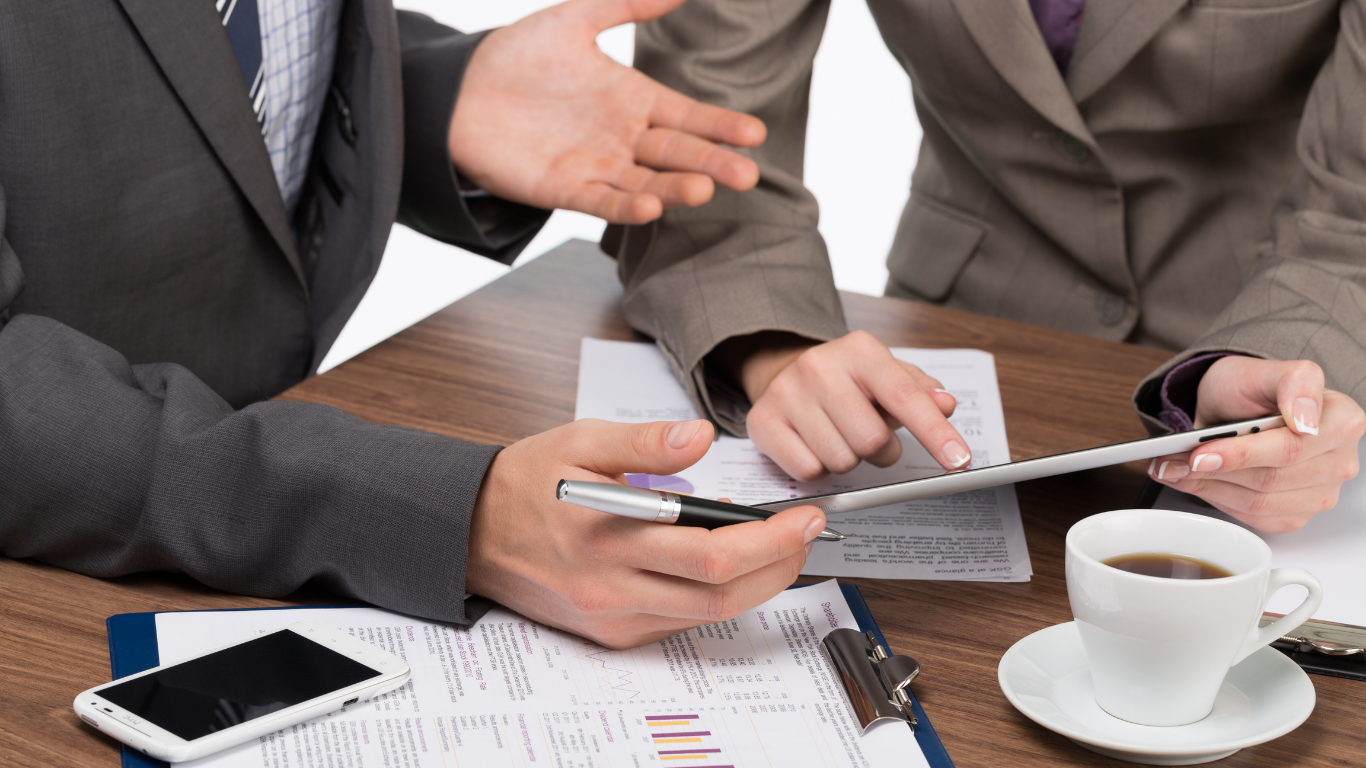 Two people at a table, discussing documents and using a tablet. Cup of coffee and a smartphone on the table.