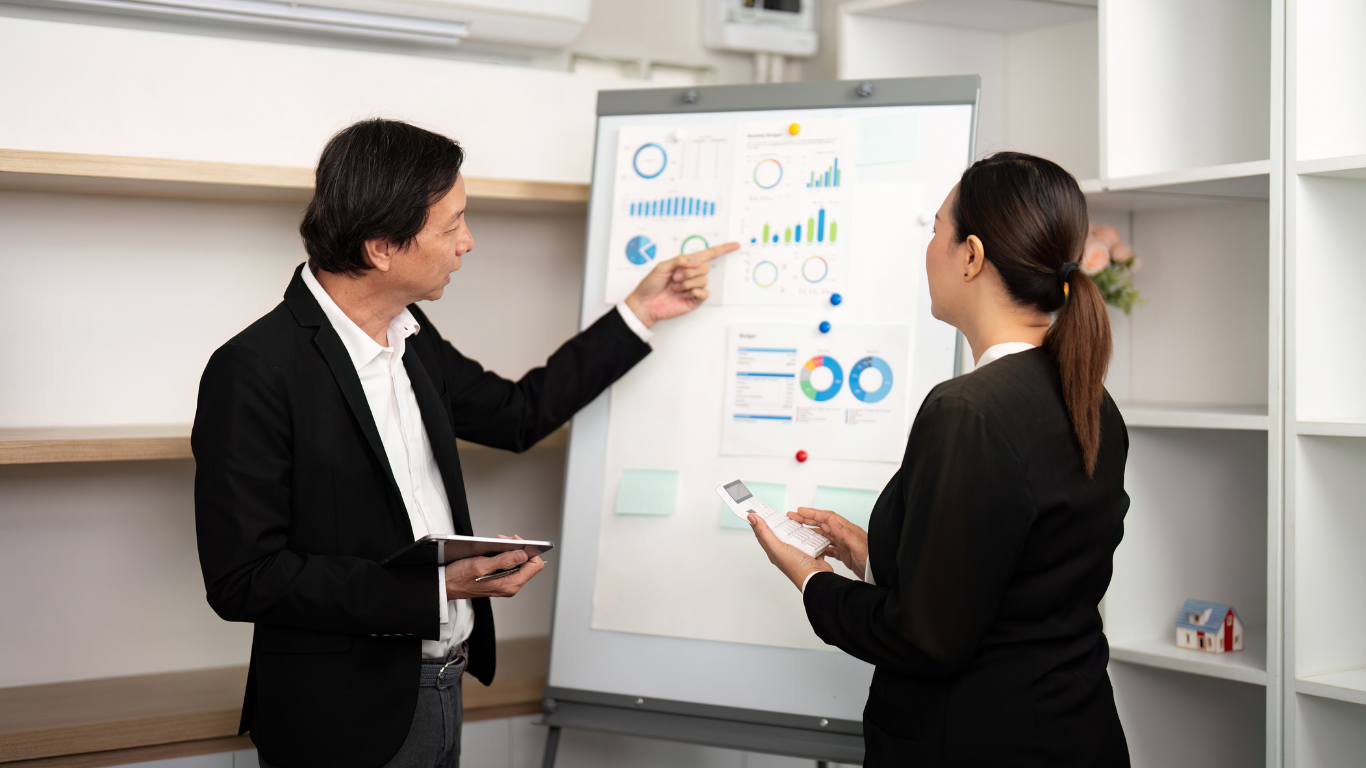Man and woman in business attire reviewing charts on a whiteboard. Man points, woman holds a phone.