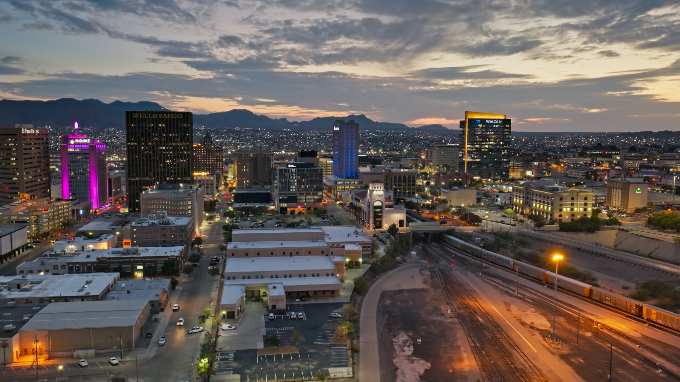 City skyline at dusk with lit buildings, mountains in the distance, and train tracks in the foreground.