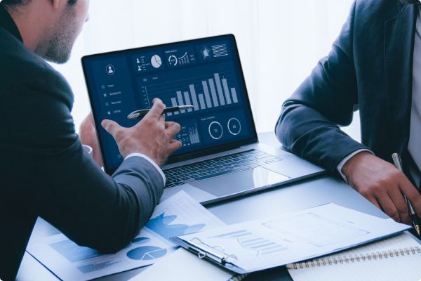 Two people in suits analyzing data on a laptop, pointing at charts and graphs in a bright office.