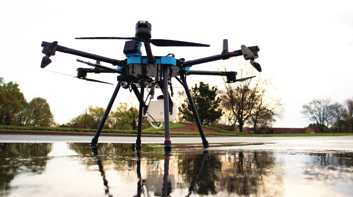 Large black-and-blue drone hovering low over a wet reflective surface in a park.
