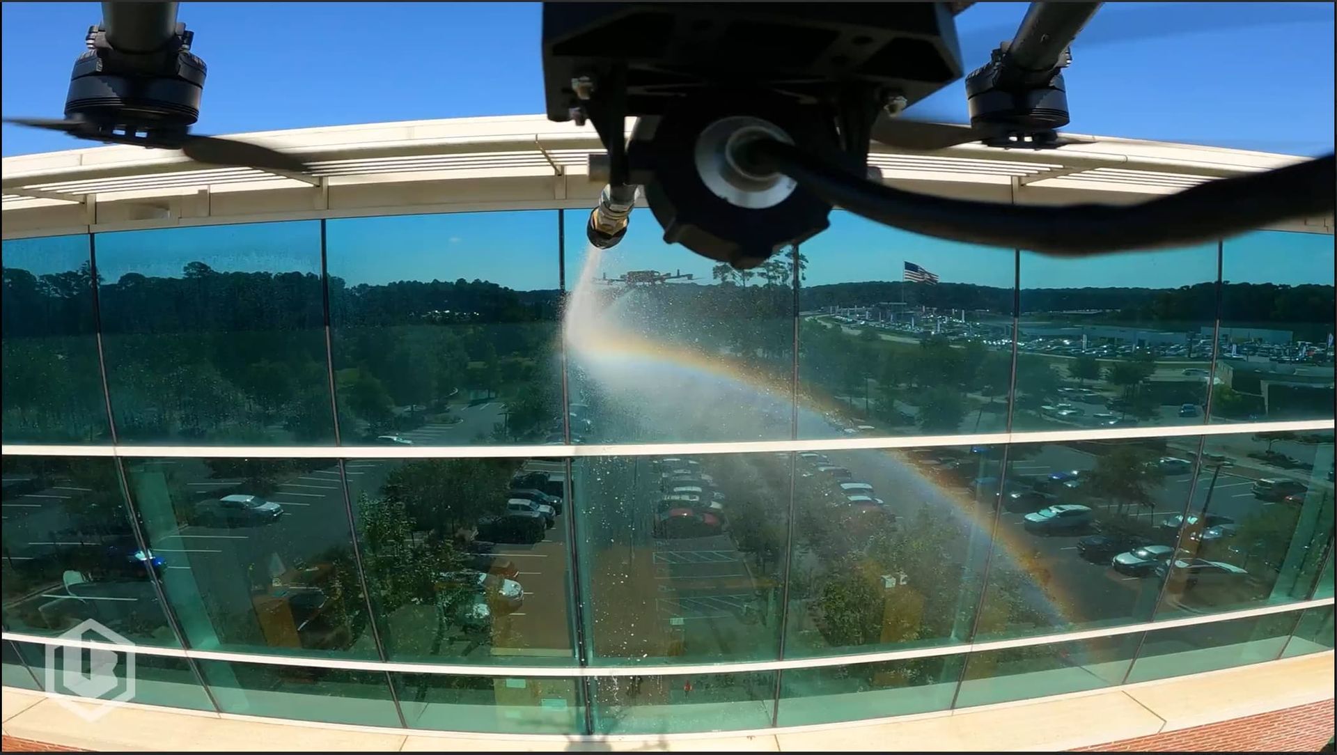 Aerial water spray on a glass-railed terrace overlooking trees and a lake under a blue sky.