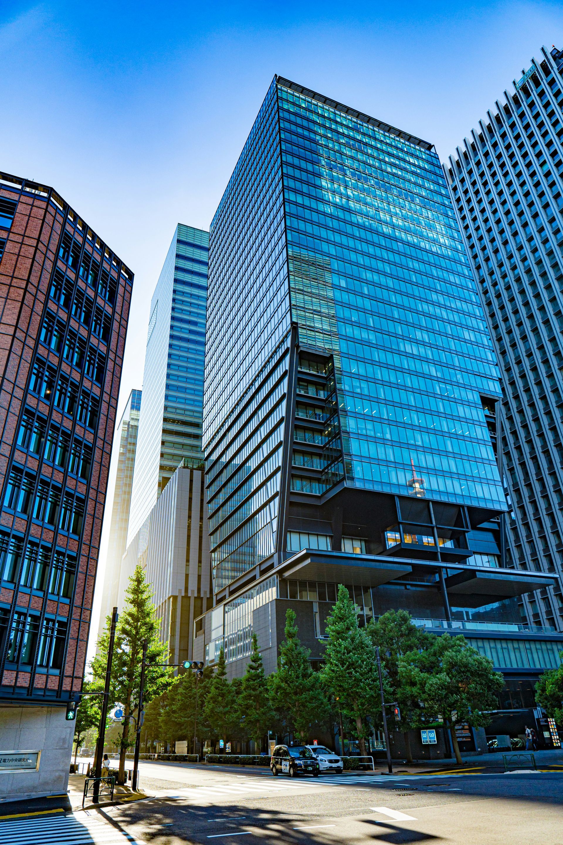 Tall blue-glass office towers on a sunny city street with trees and cars below