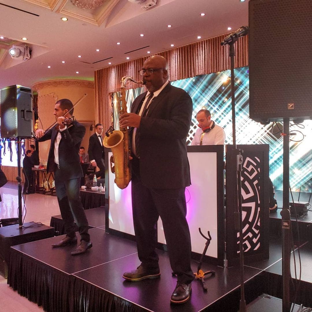 Corporate Events, Band performing on stage: saxophonist, flutist, and DJ playing at an event. Stage is decorated with lights.