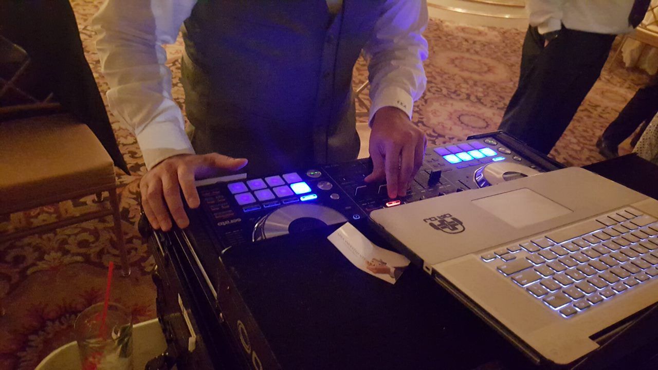 DJ mixing music at an event, operating a soundboard with laptop, in a room with a patterned carpet.