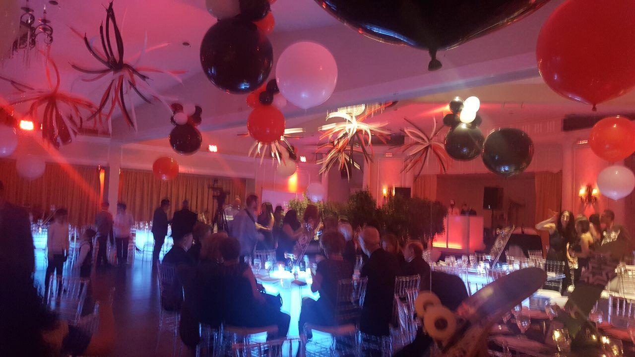 Party scene with guests seated at tables, red, white, and black balloons, and decorative lighting.