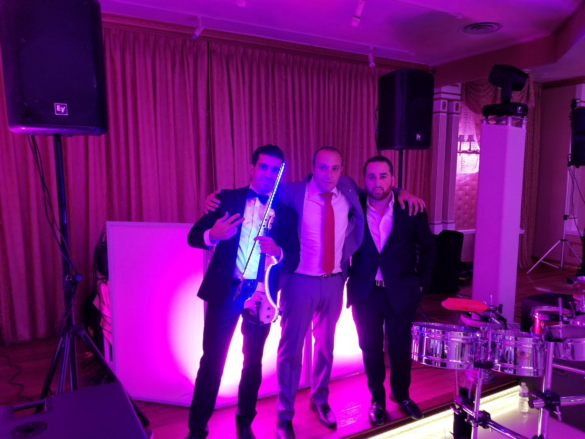 Three men pose with a DJ setup, lit with purple light. One holds a glowing instrument. Formal attire.