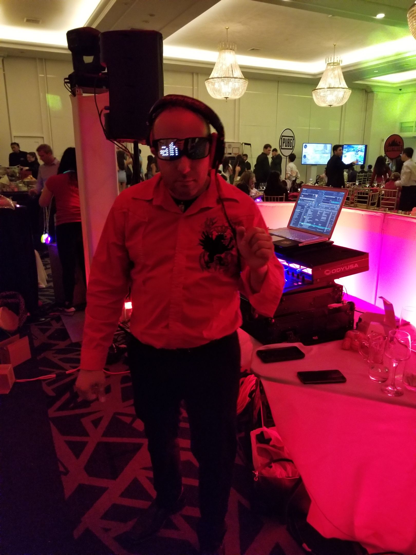 Man in red shirt and glasses at DJ setup, lit with red light.