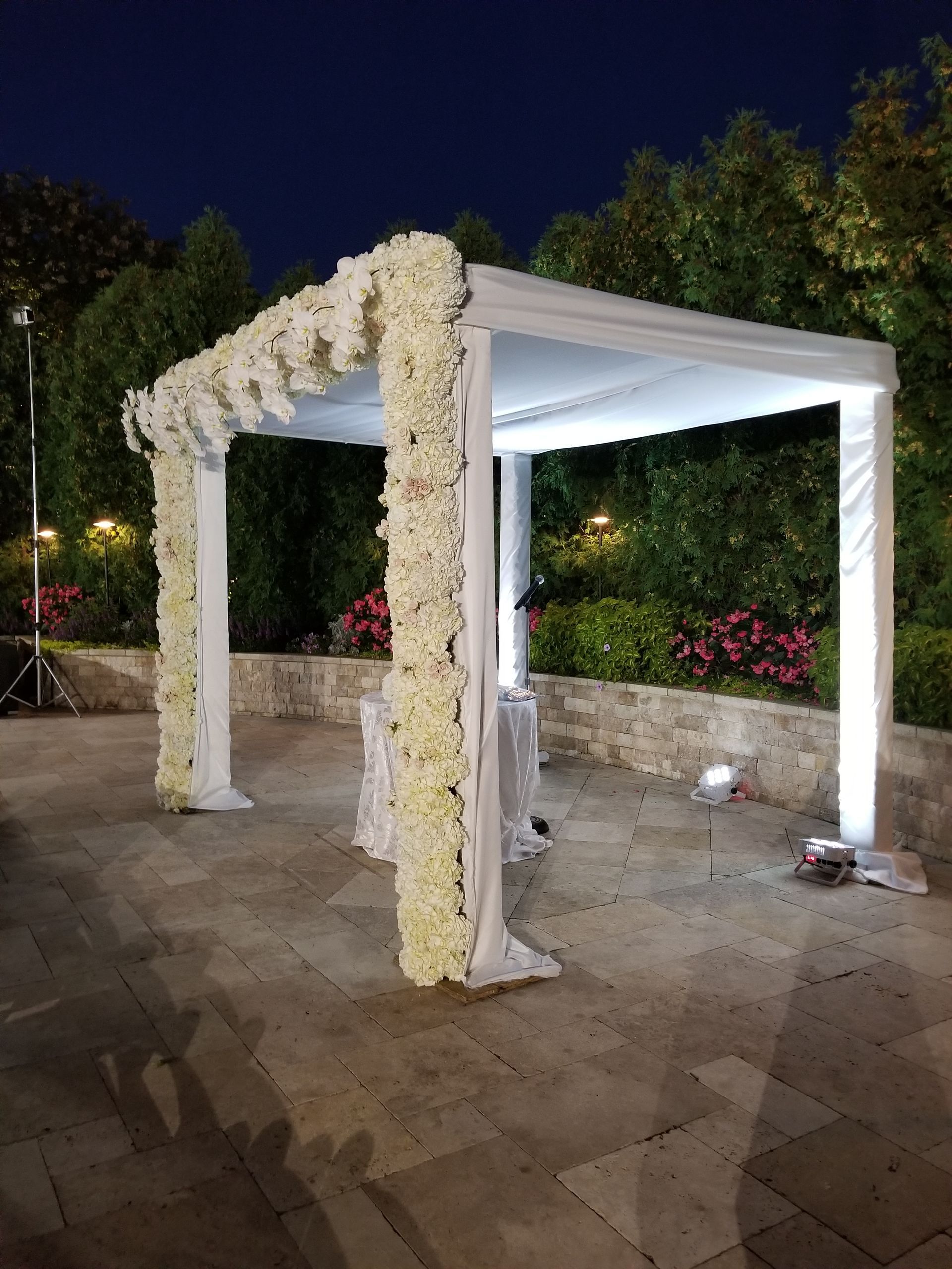 White floral-covered chuppah outdoors at night, with draped fabric and surrounding greenery.