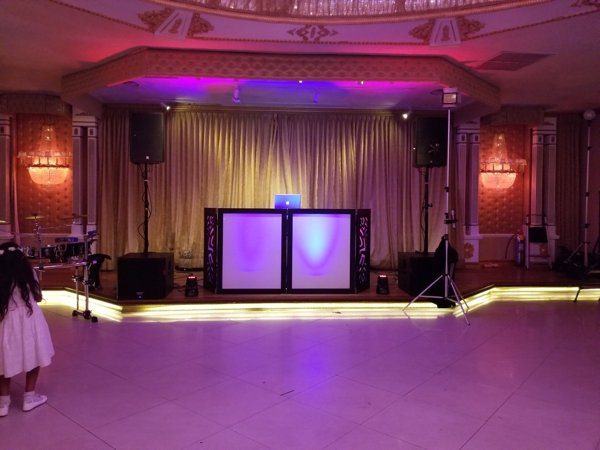 DJ setup on stage, lit with purple and gold, in a ballroom with ornate columns and a young girl in white.