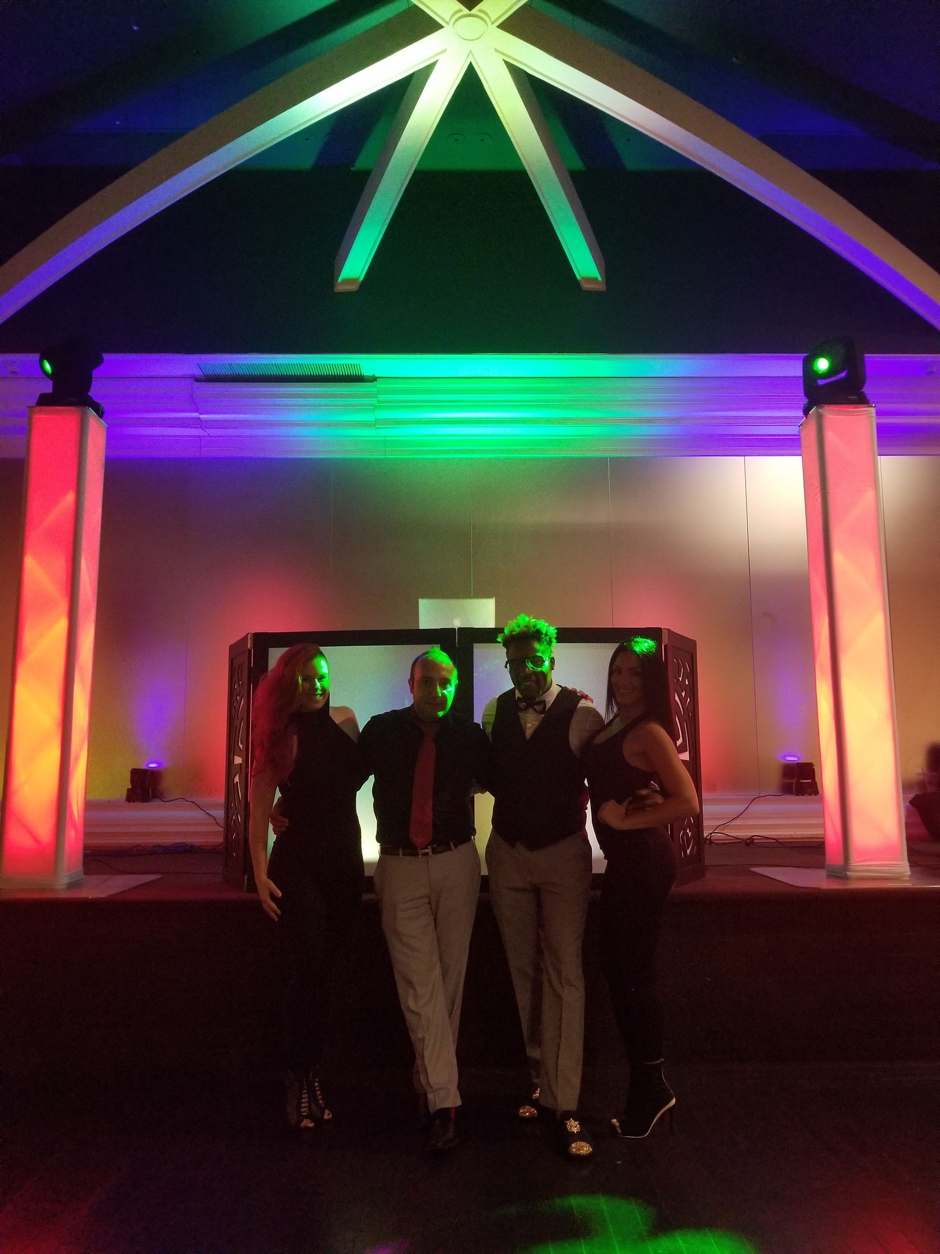 Four people pose on a stage, lit with colorful lights. The group is in a performance setting, possibly a party or show.