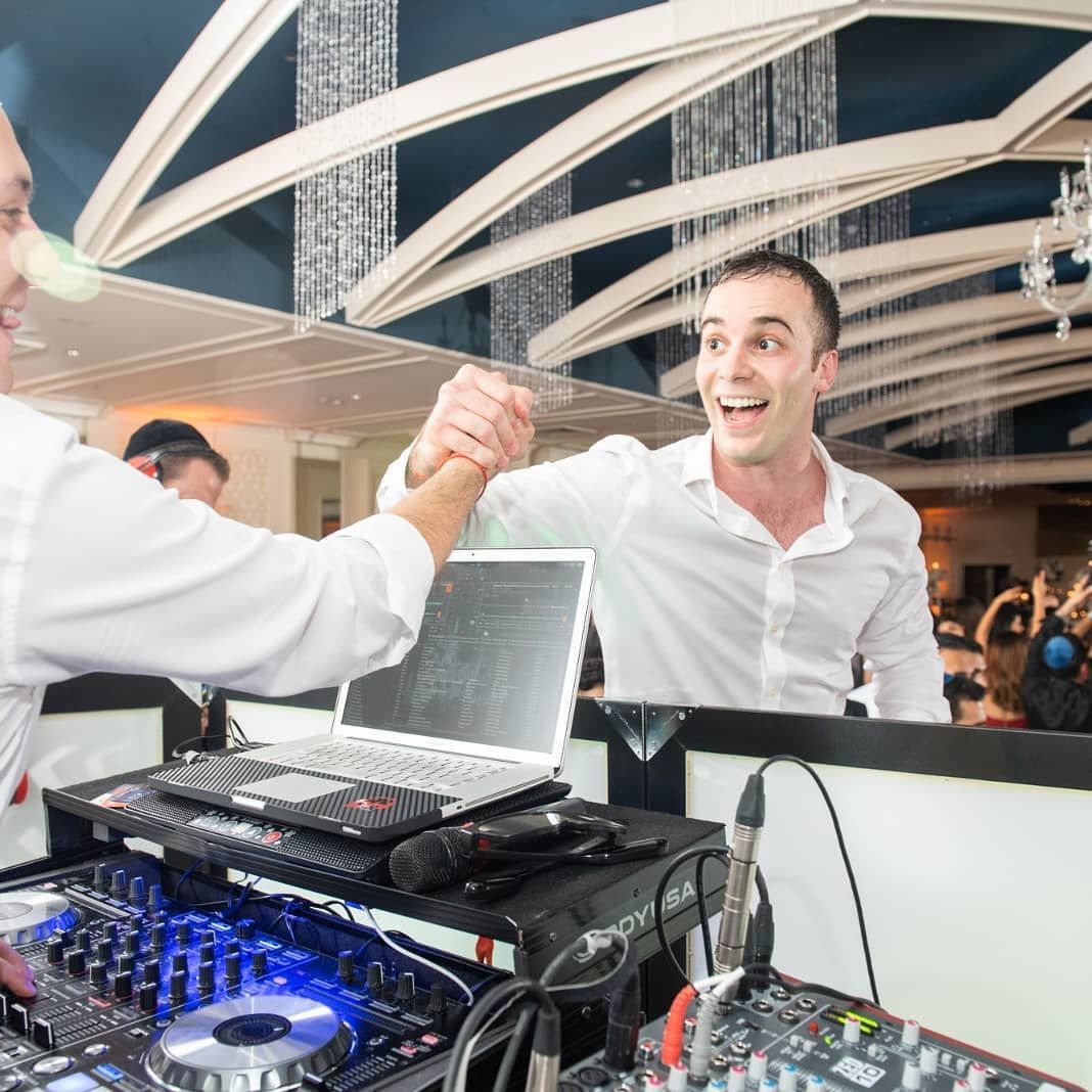 Two men high-fiving over DJ equipment at a party. One man smiles, expressing excitement.