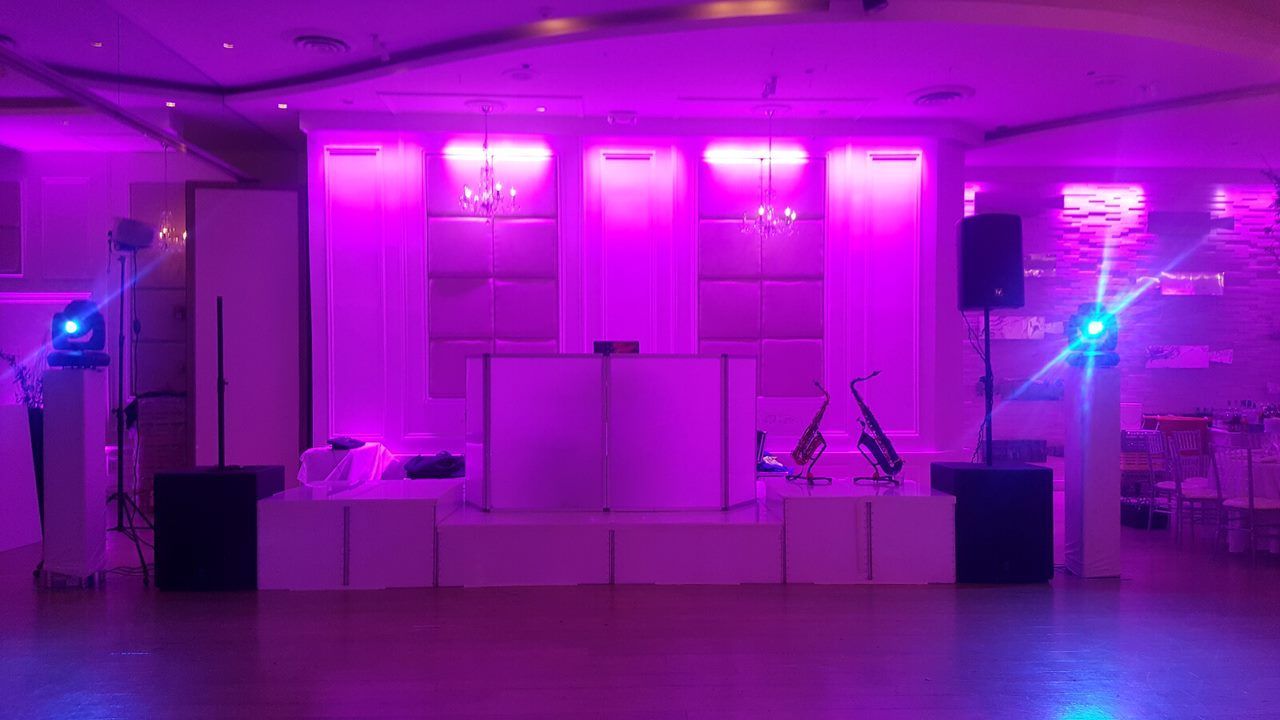 A stage setup with white risers, DJ booth, speakers, and purple uplighting in an event space.