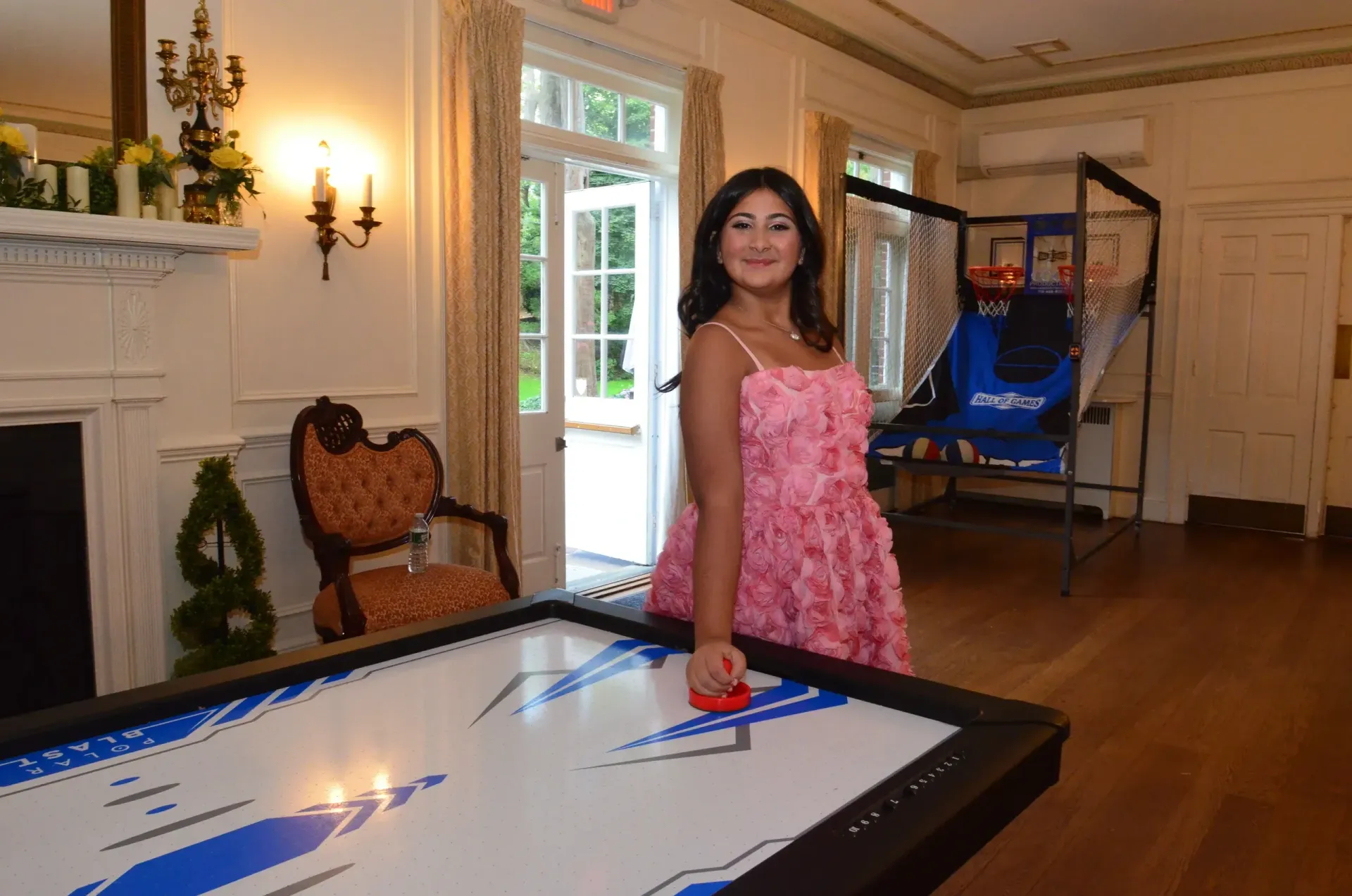 Woman in pink dress plays air hockey in a room with a basketball game and fireplace.