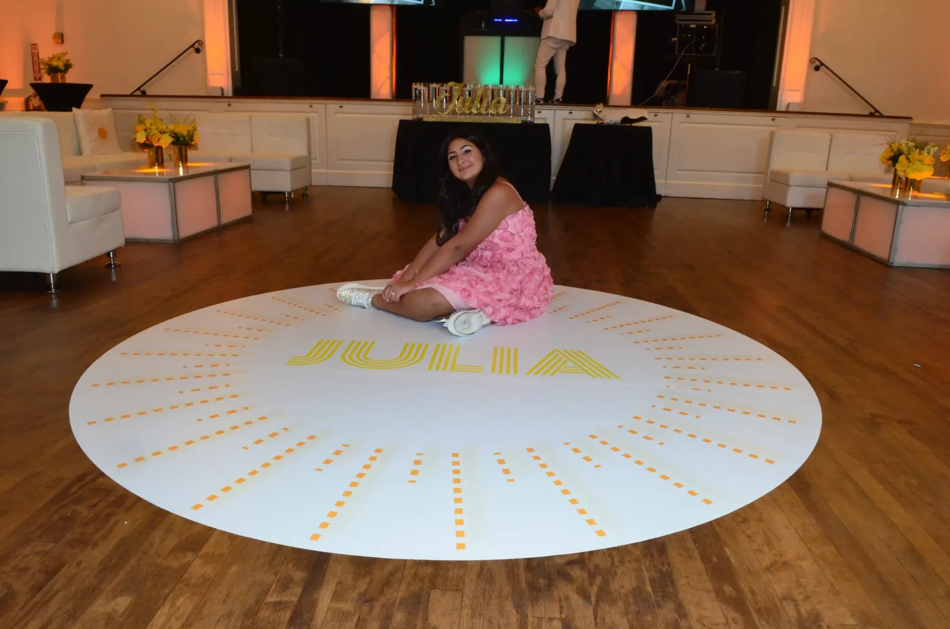 Person sitting on white dance floor with yellow design; stage and seating in the background.
