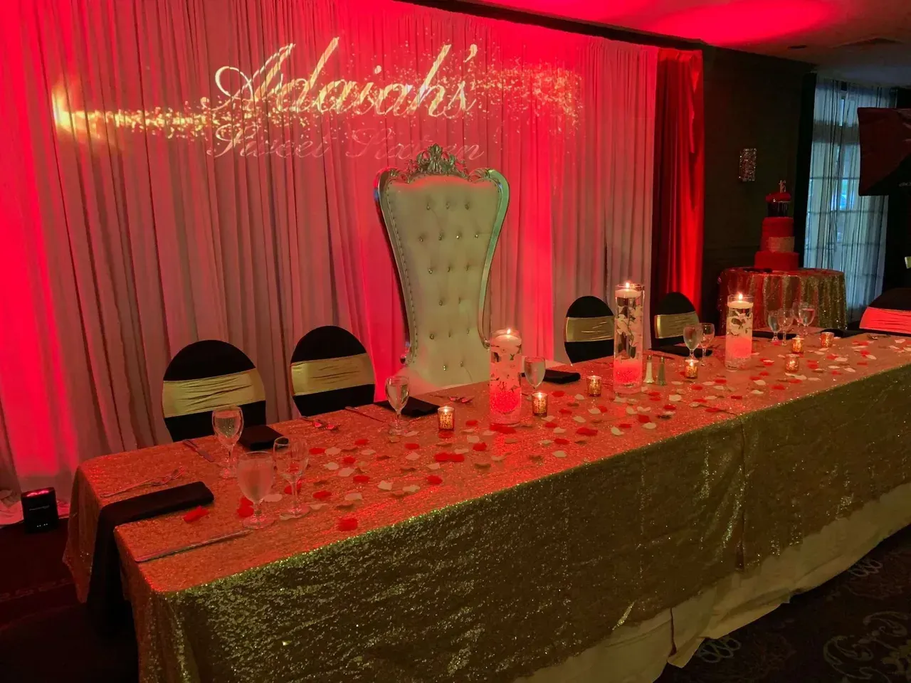 A decorated table with a gold sequin tablecloth, large white chair, and red and gold accents for an event.