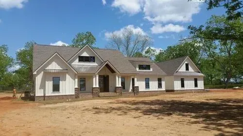 A large white house with a gray roof is sitting in the middle of a dirt field.