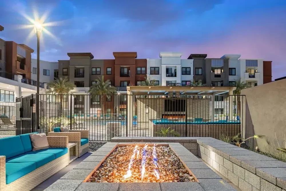 Outdoor fire pit and turquoise seating in front of apartment building with pool and evening sky.
