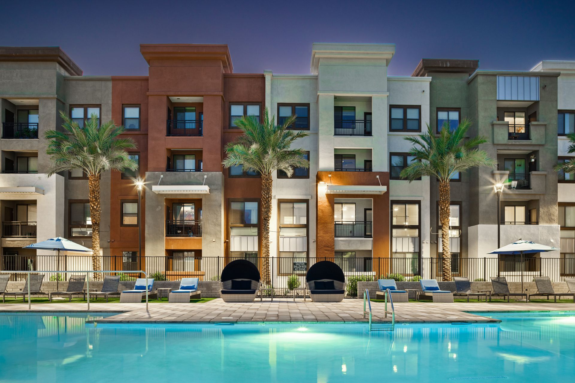 Modern apartment complex with balconies and palm trees reflected in a turquoise pool at dusk