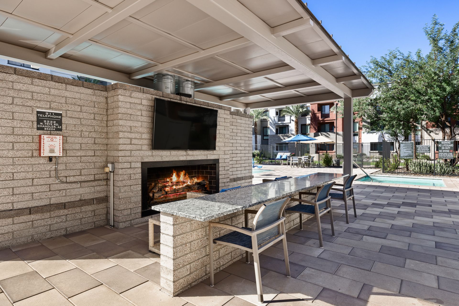 Outdoor poolside patio with a stone fireplace, covered seating, and tables near an apartment complex