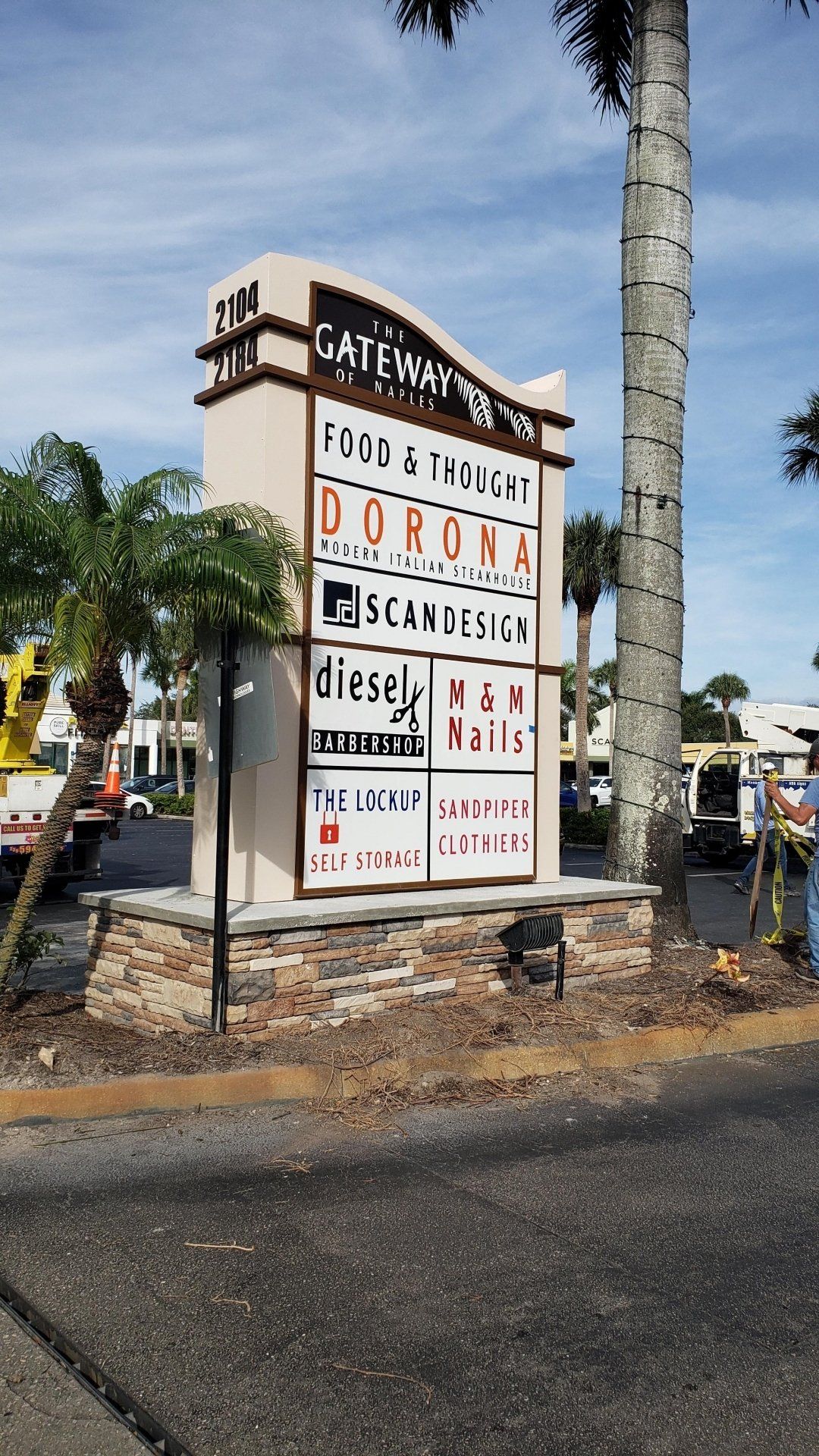A large sign is sitting on the side of the road next to a palm tree.