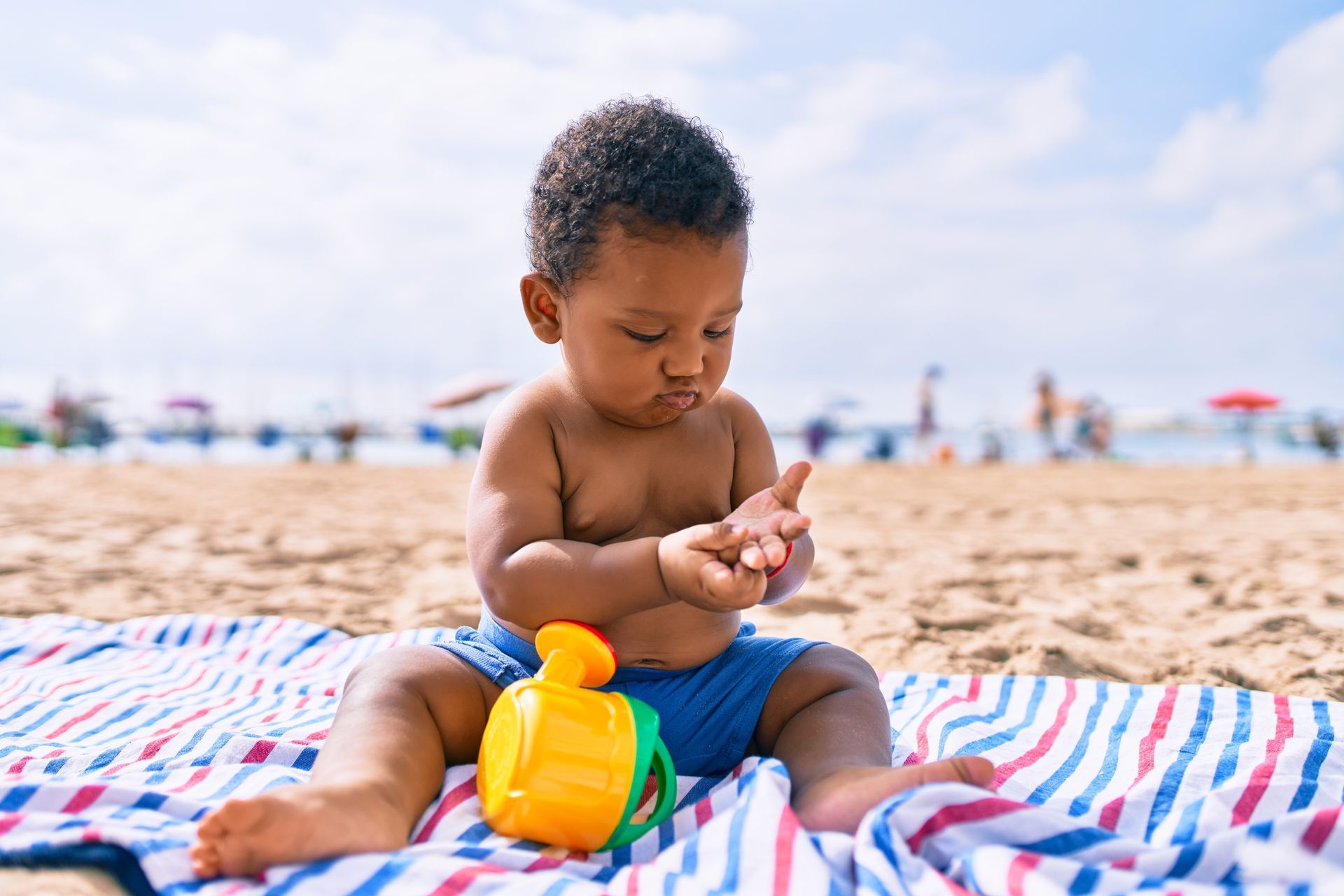 Baby sitting on a beach towel, playing with a toy.