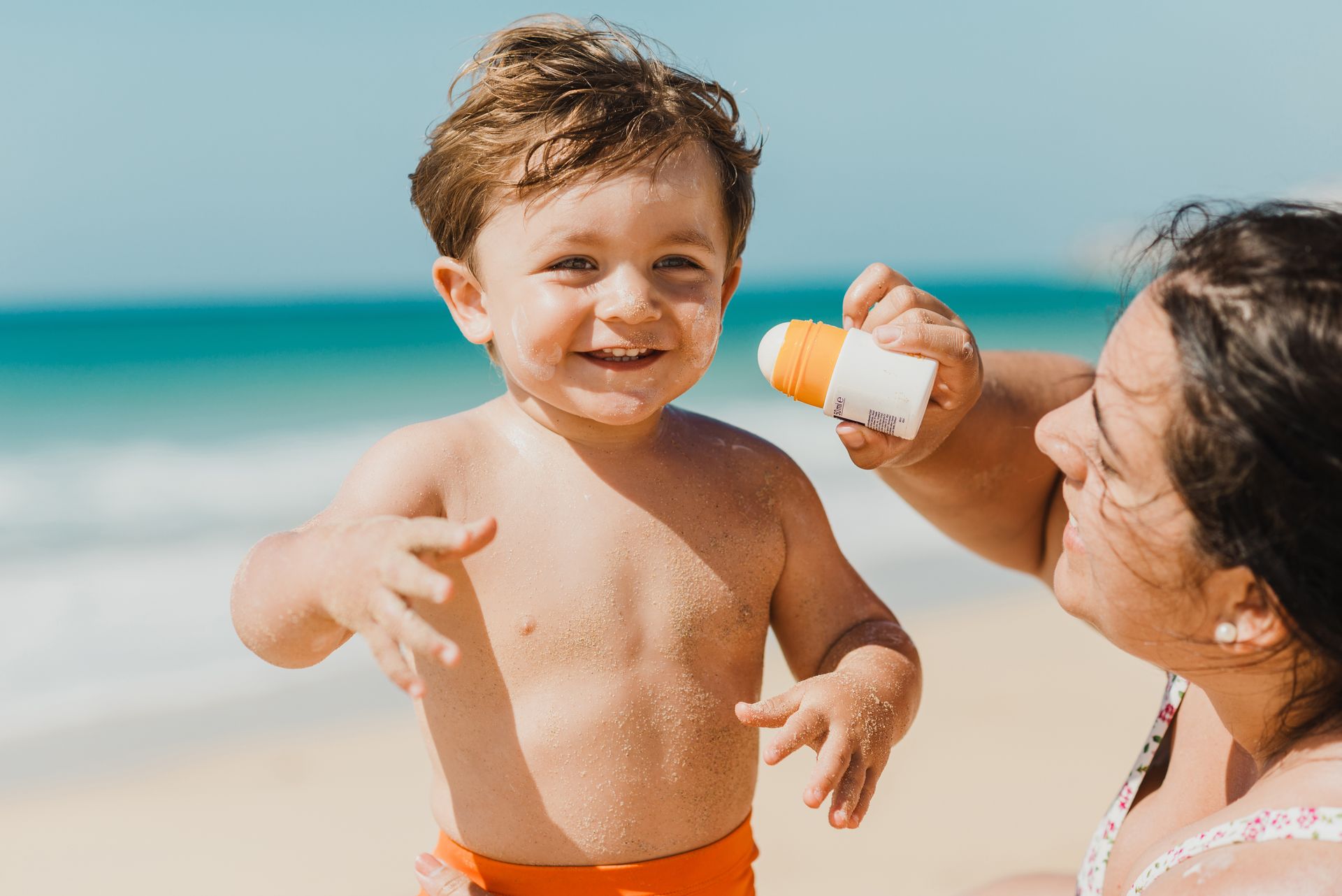 A woman is applying sunscreen to a young boy on the beach.