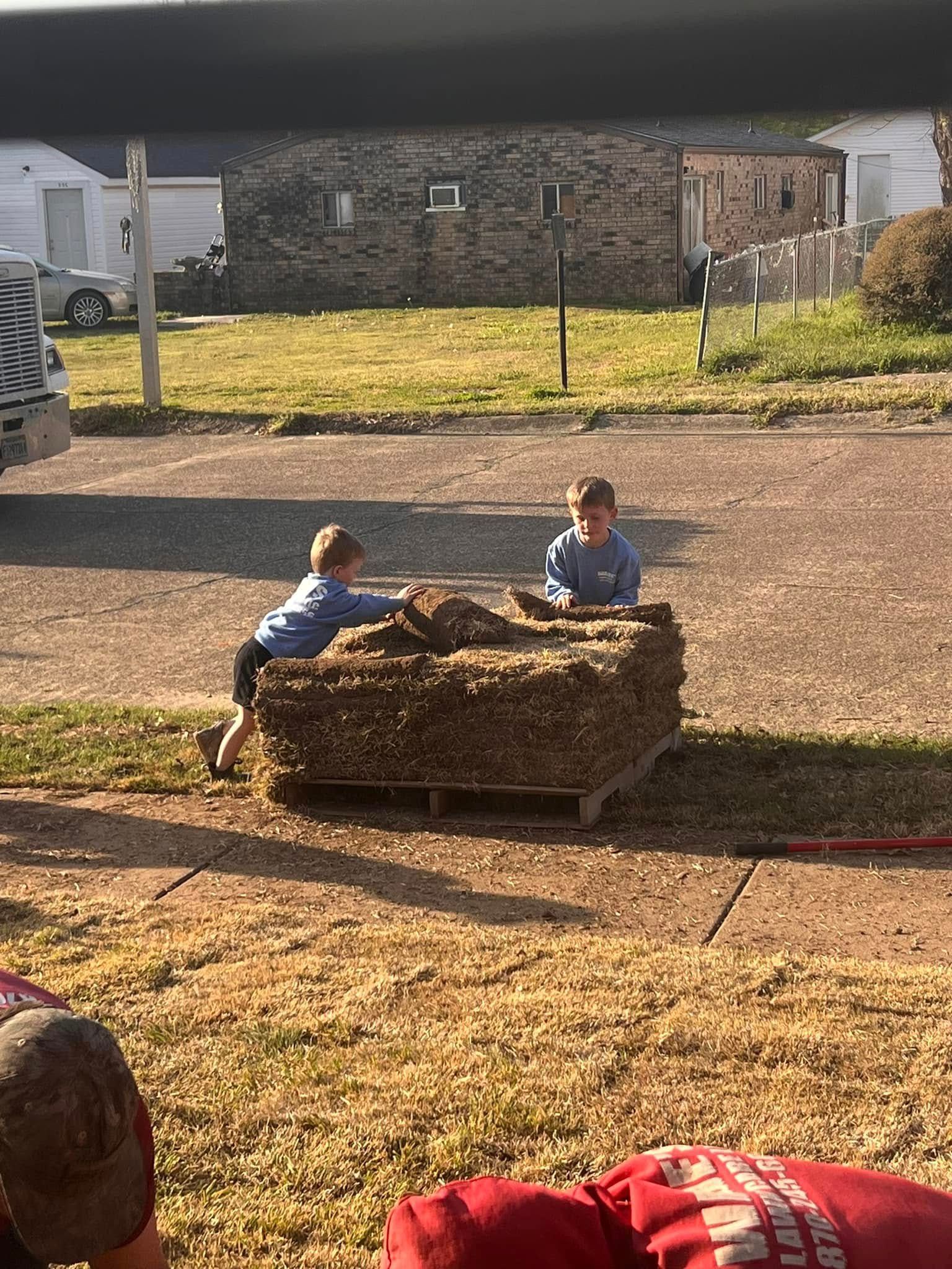 Two children playing on a hay bale in a sunny outdoor yard