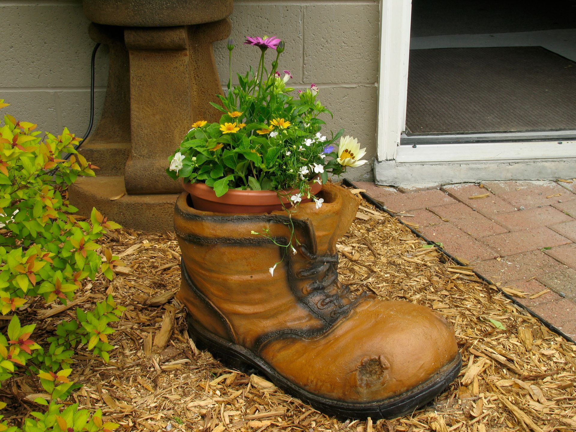Brown boot planter filled with flowers, set on wood chips near a building entrance.