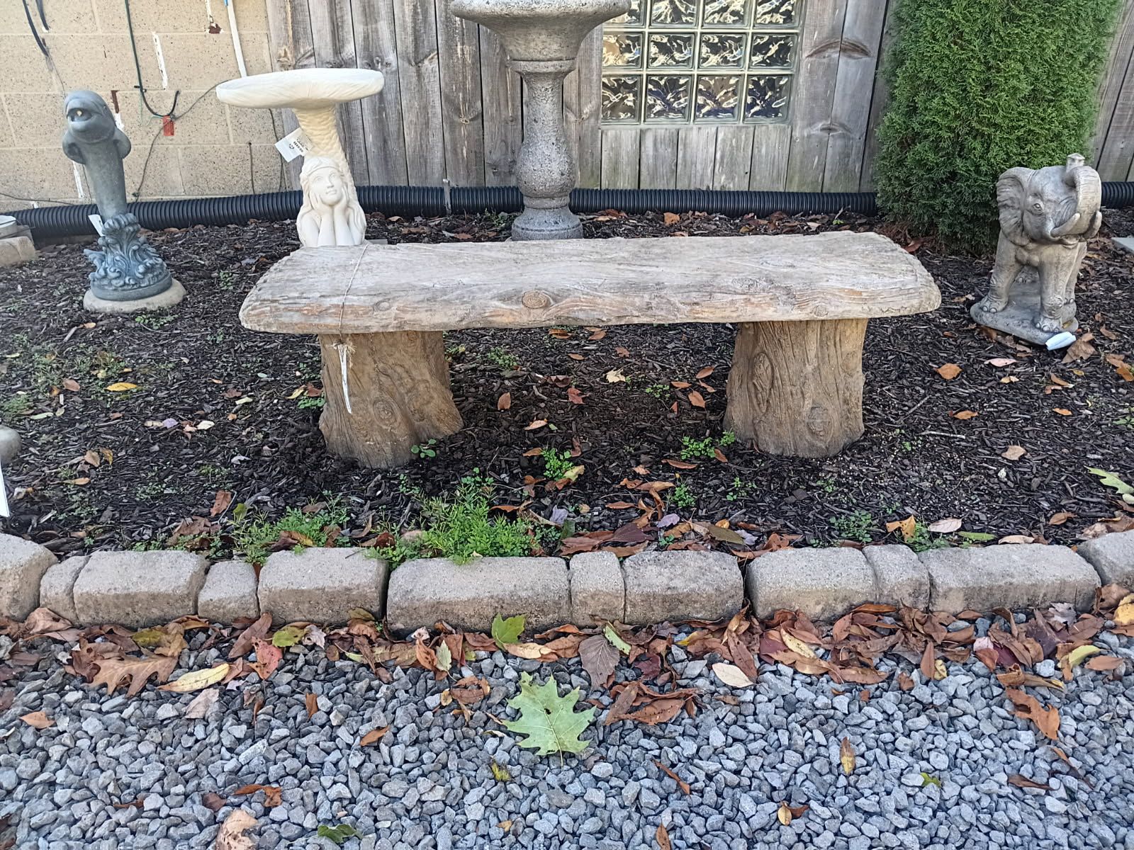 Stone bench in a garden setting, surrounded by other garden decor and a brick border.