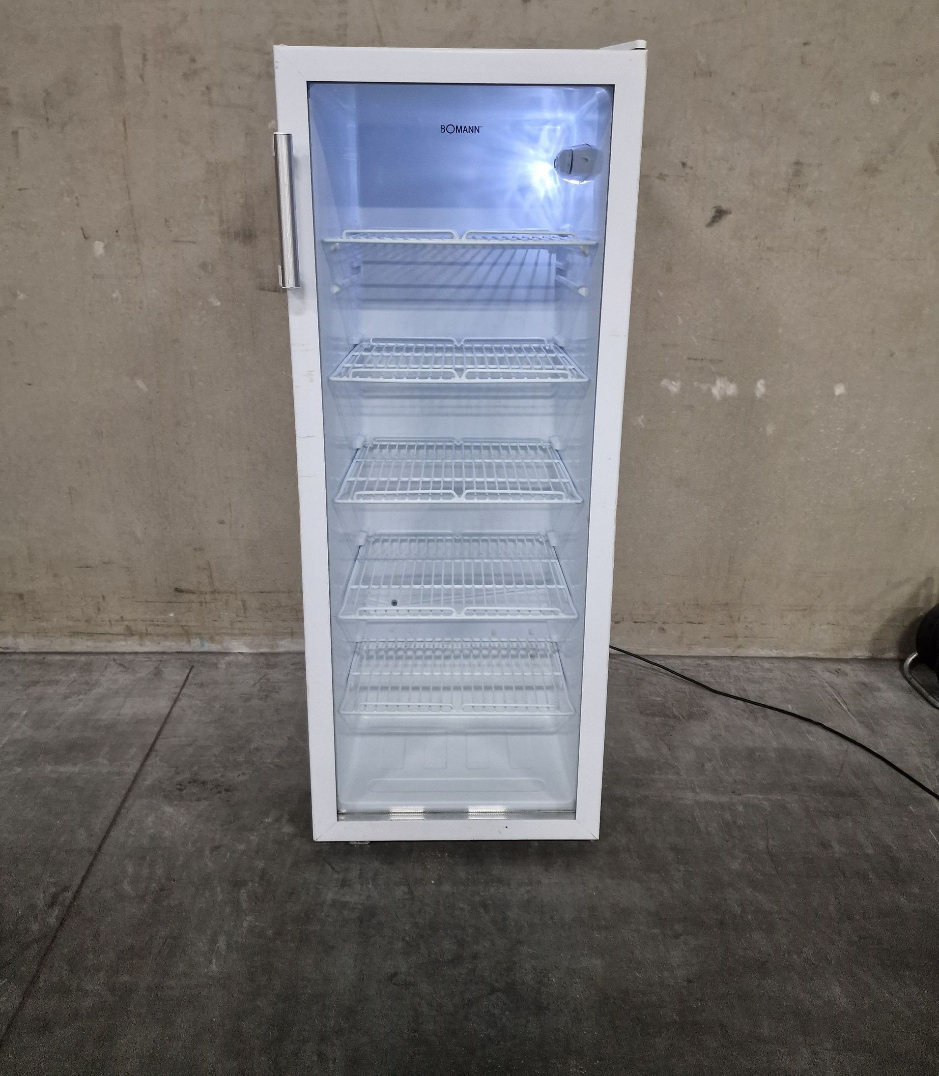 Een witte koelkast met een glazen deur staat op een betonnen vloer.