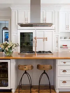 A kitchen with white cabinets , wooden counter tops , stools and a refrigerator.