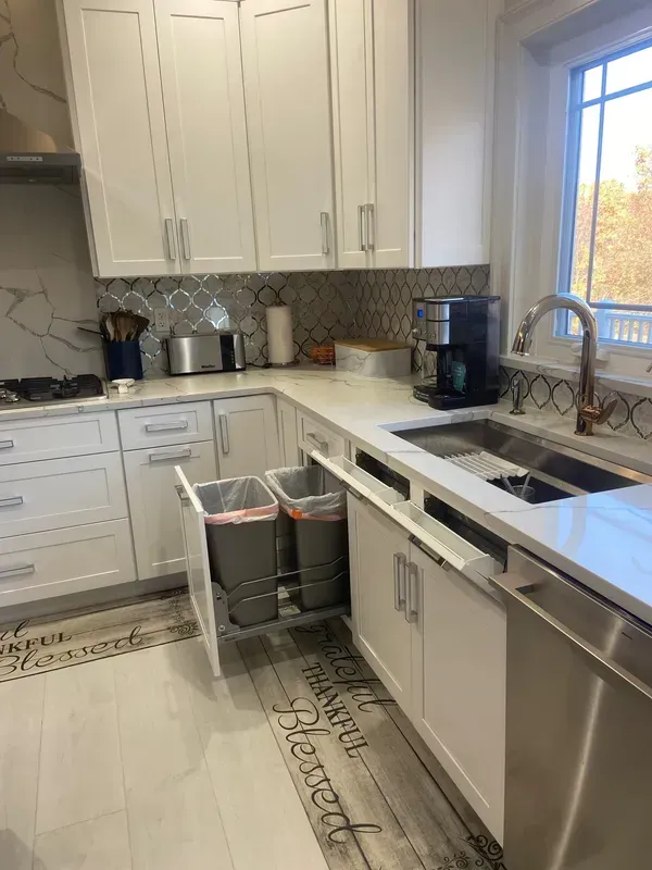 A kitchen with white cabinets , stainless steel appliances , a sink , and a trash can.