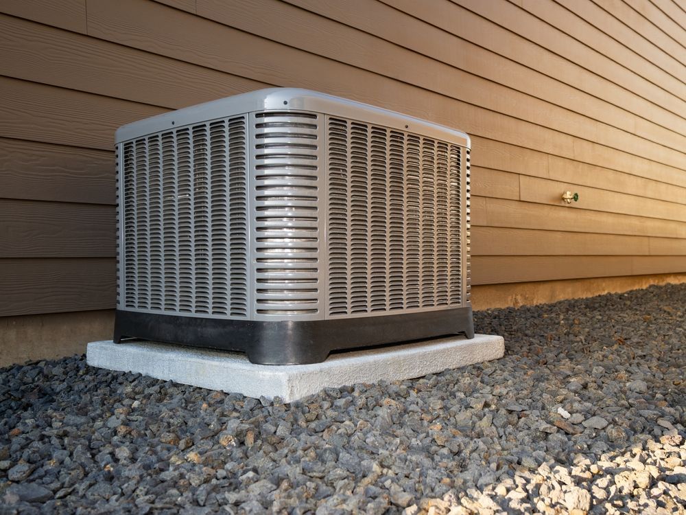 An air conditioner is sitting outside of a house on gravel.