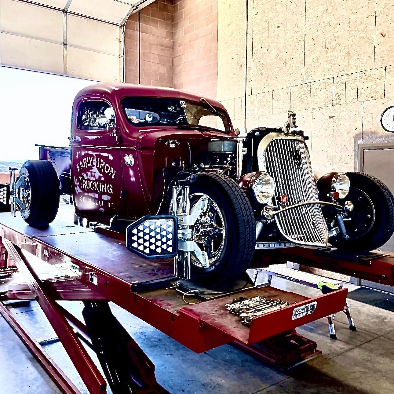 A burgundy hot rod truck on a red lift undergoing wheel alignment in a garage.