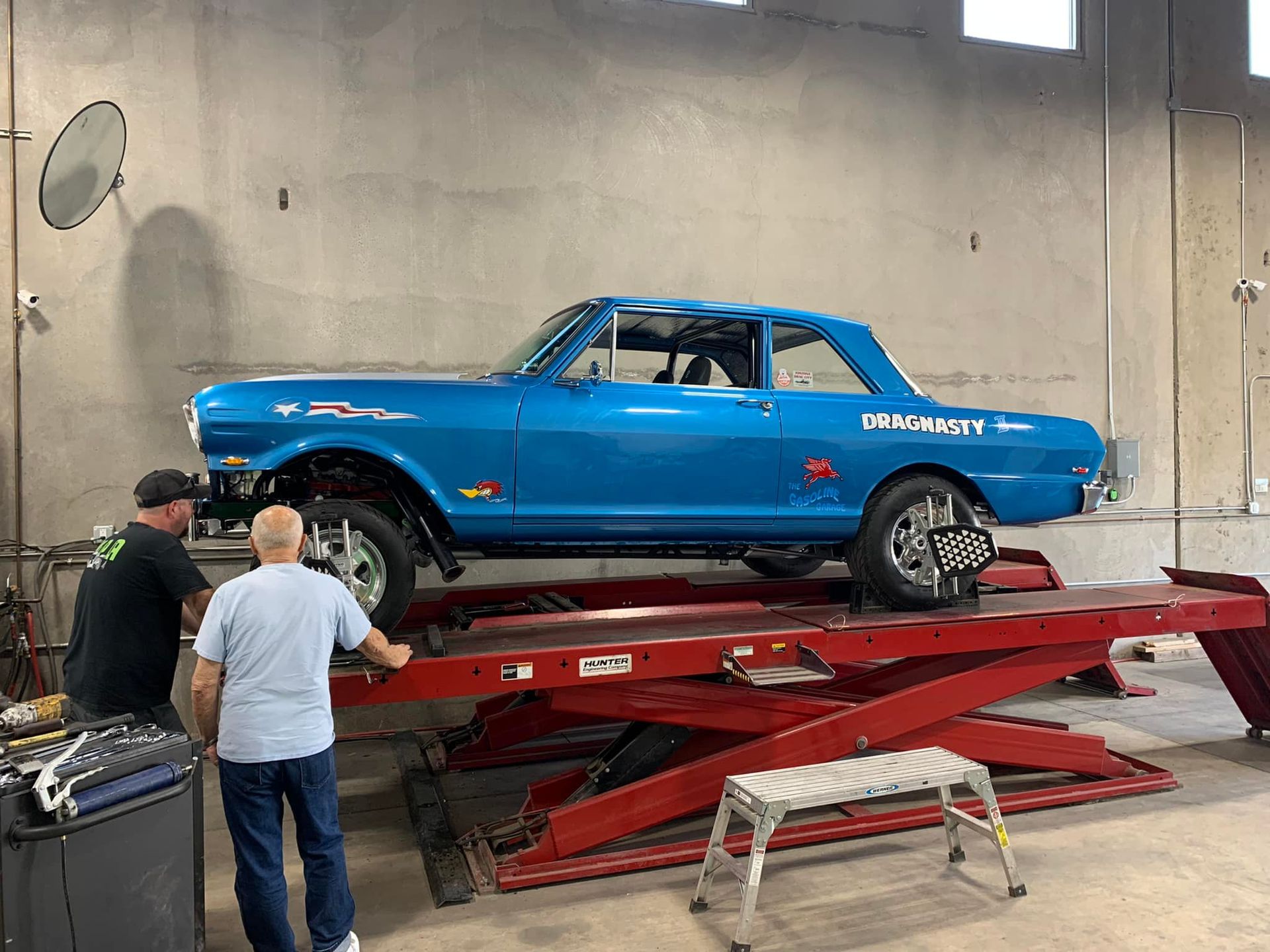 Two men inspect a blue classic car on a red hydraulic lift inside a garage.
