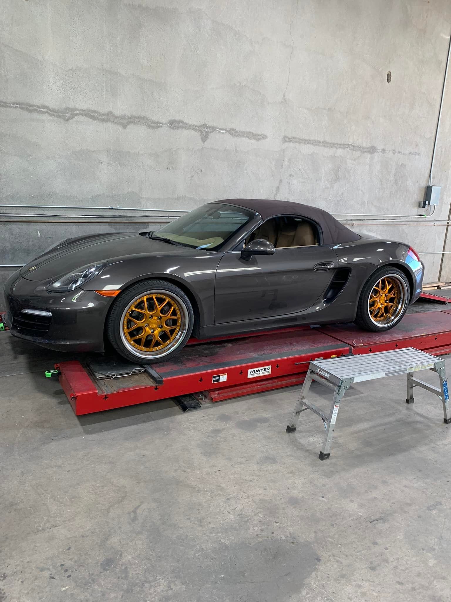 Dark grey Porsche Boxster on a lift, gold rims, top down, inside a garage.