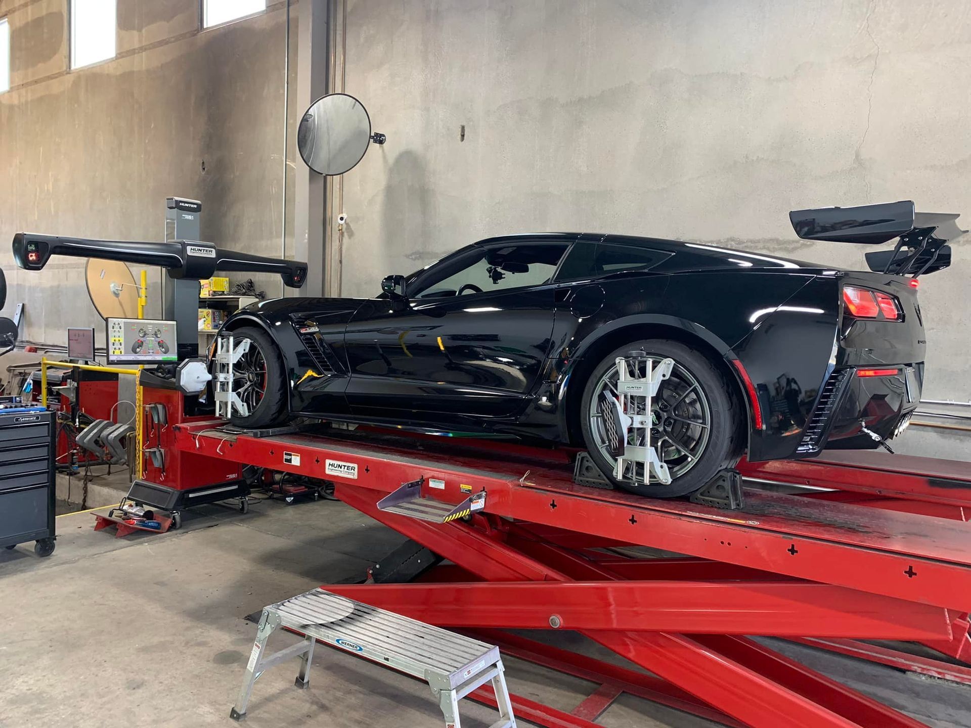 Black sports car on a red lift undergoing wheel alignment in a garage.