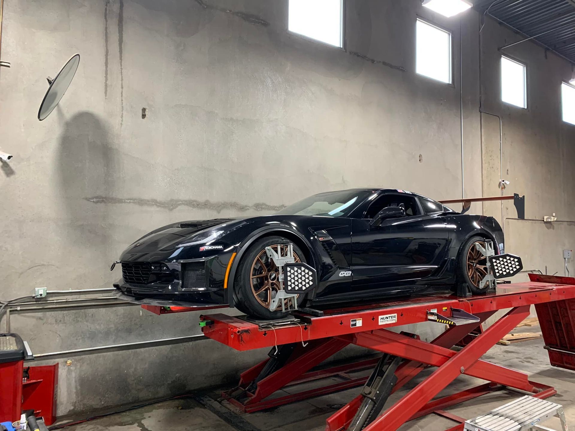 Black Corvette on a red lift in a garage, undergoing a wheel alignment.