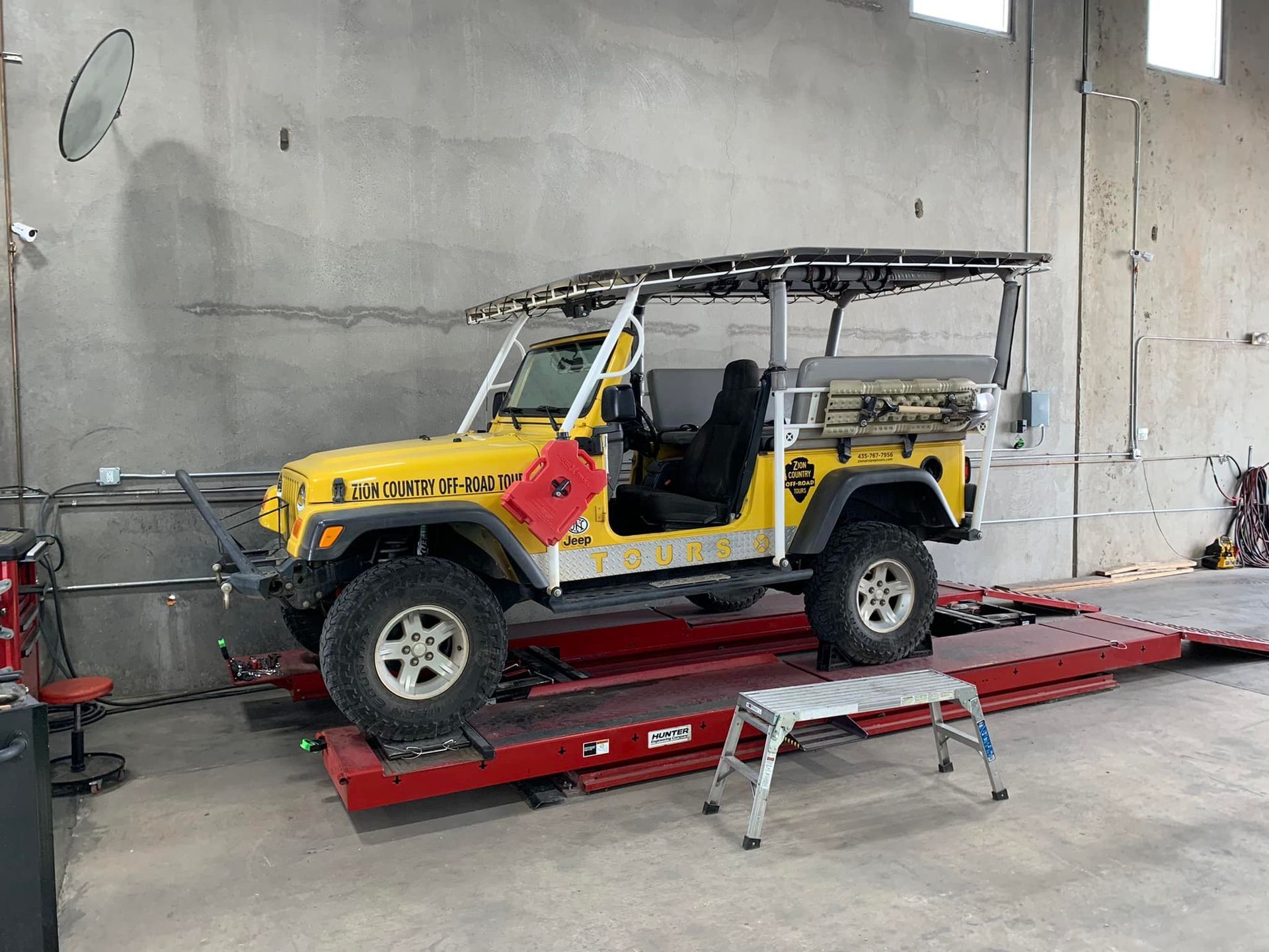 Yellow Jeep on a red lift inside a garage, with a roof rack and step stool in front.
