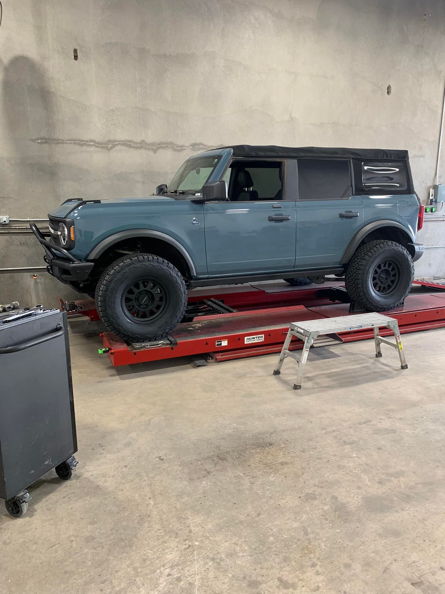 Blue Ford Bronco SUV on a red car lift in a garage. Black wheels, small step stool nearby.