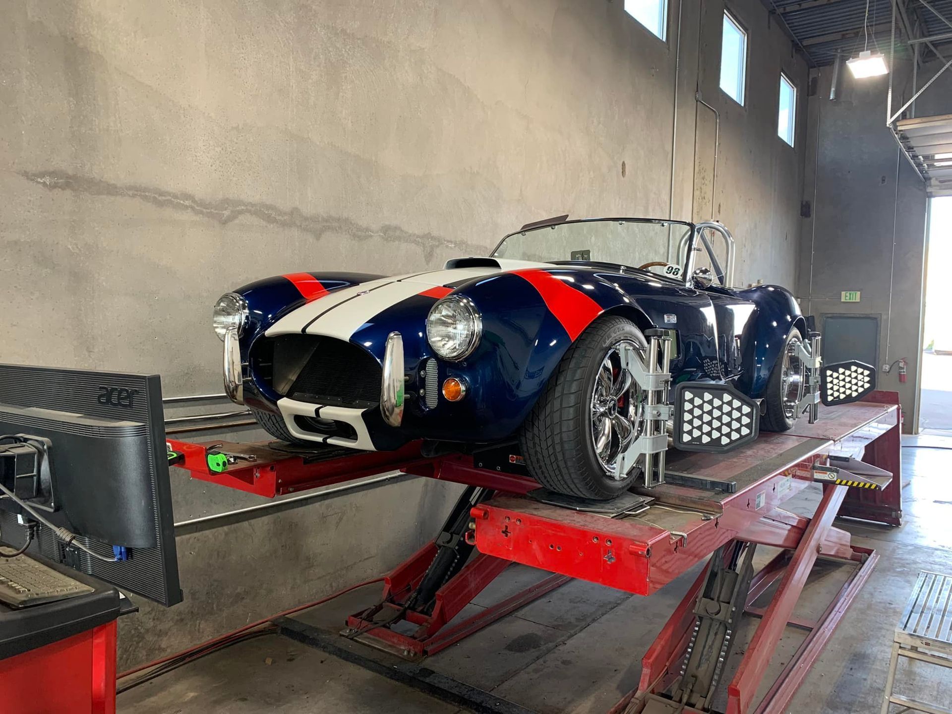 Blue and white Shelby Cobra roadster on a red lift undergoing wheel alignment in a garage.