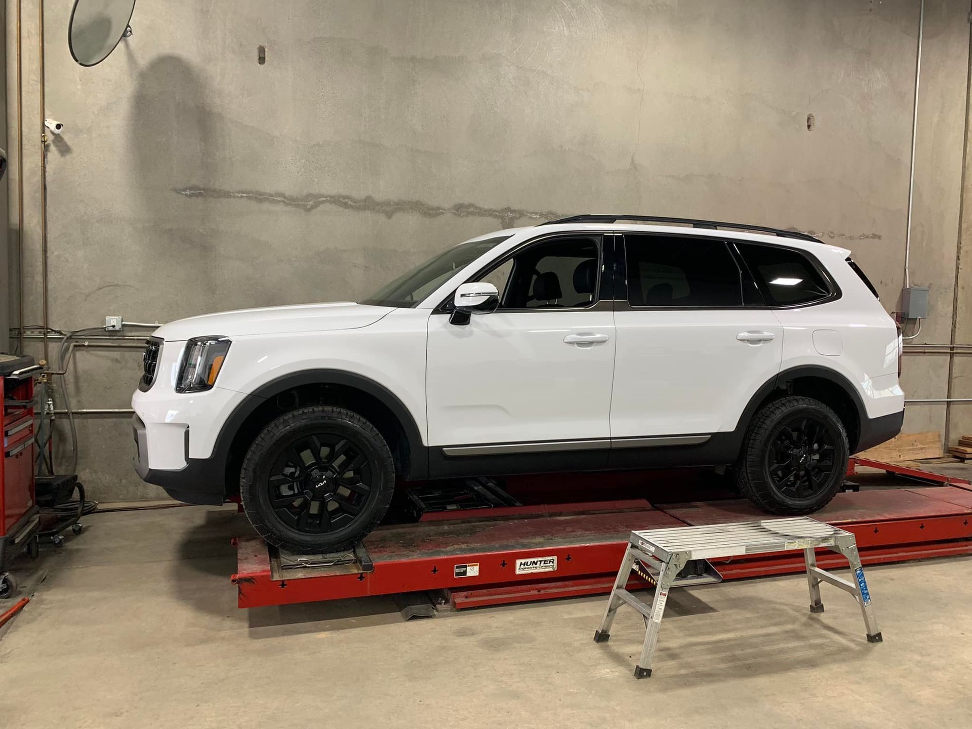 White SUV on a red lift in a garage, black wheels, against a gray wall, step stool in front.