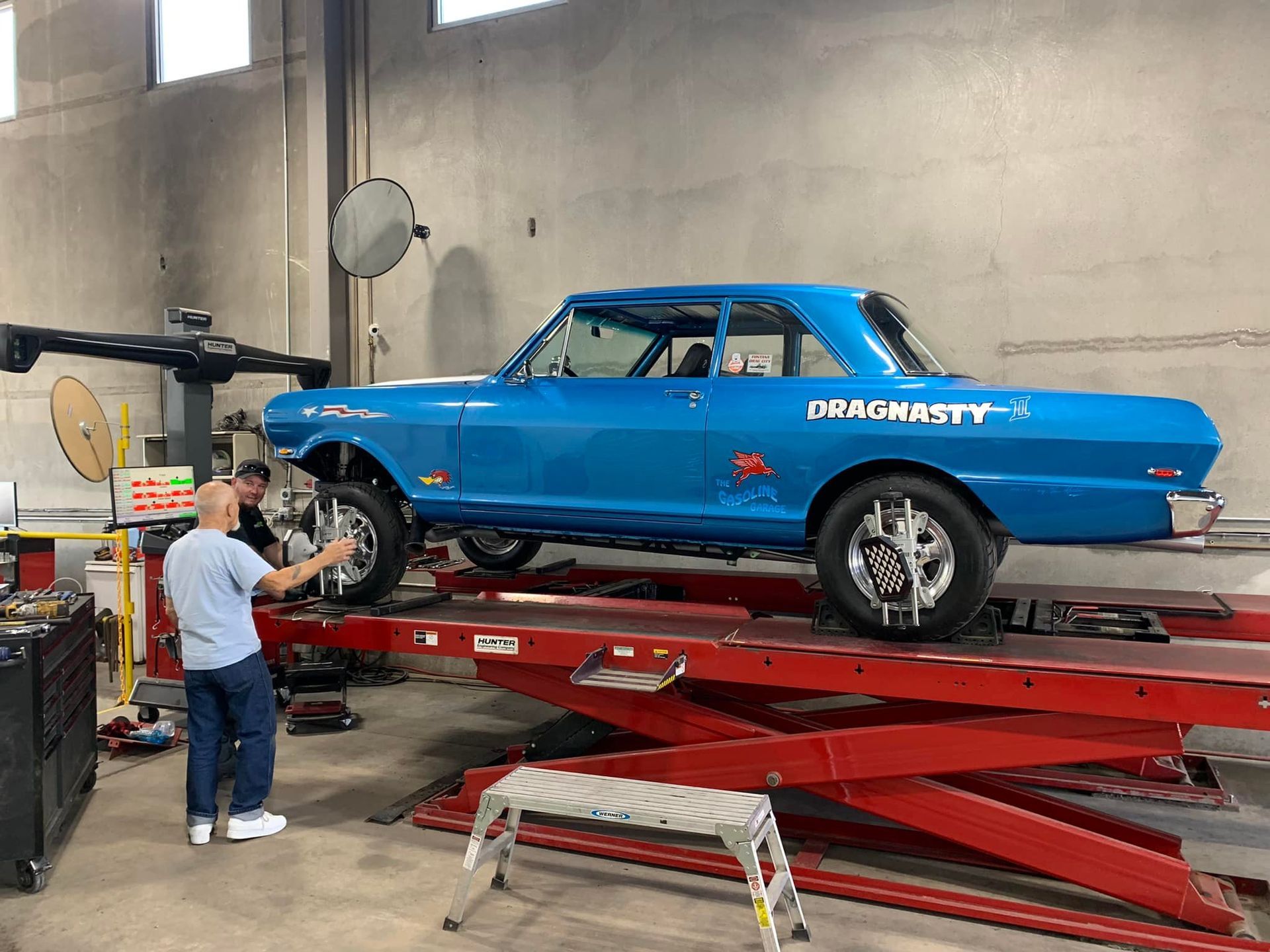 Blue classic car on a lift in a garage, two men working.