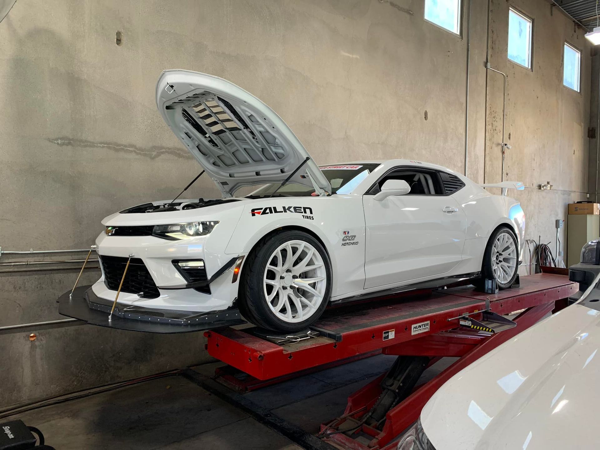 White Chevrolet Camaro on a red hydraulic lift with hood open, in a garage.
