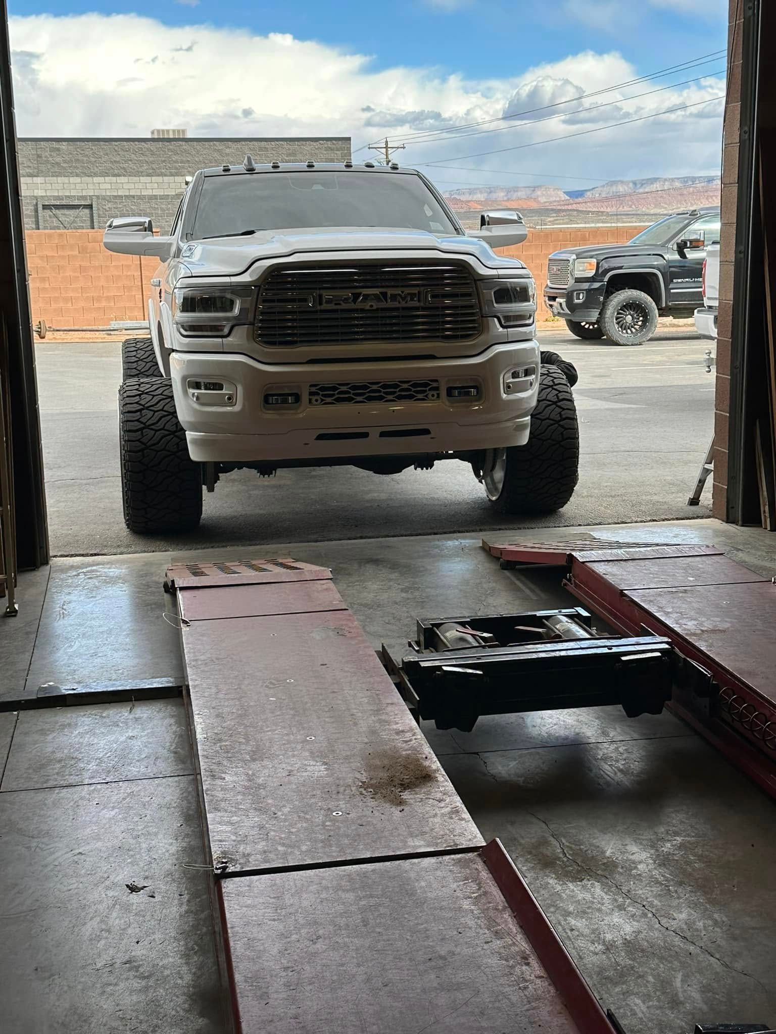 Large white truck on a lift inside a garage. Another truck is visible in the background. Blue sky overhead.